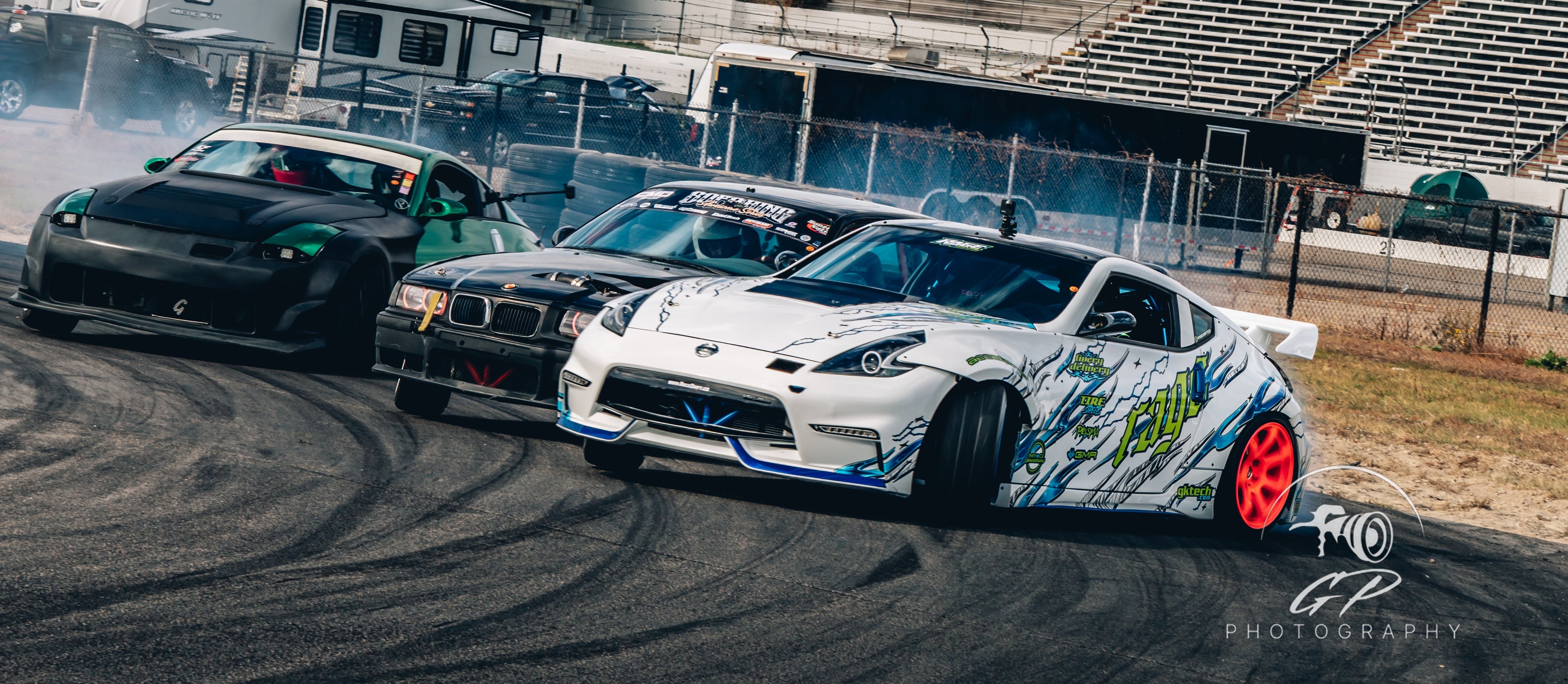 Three cars racing on a track with a cloudy sky and empty stands in the background.