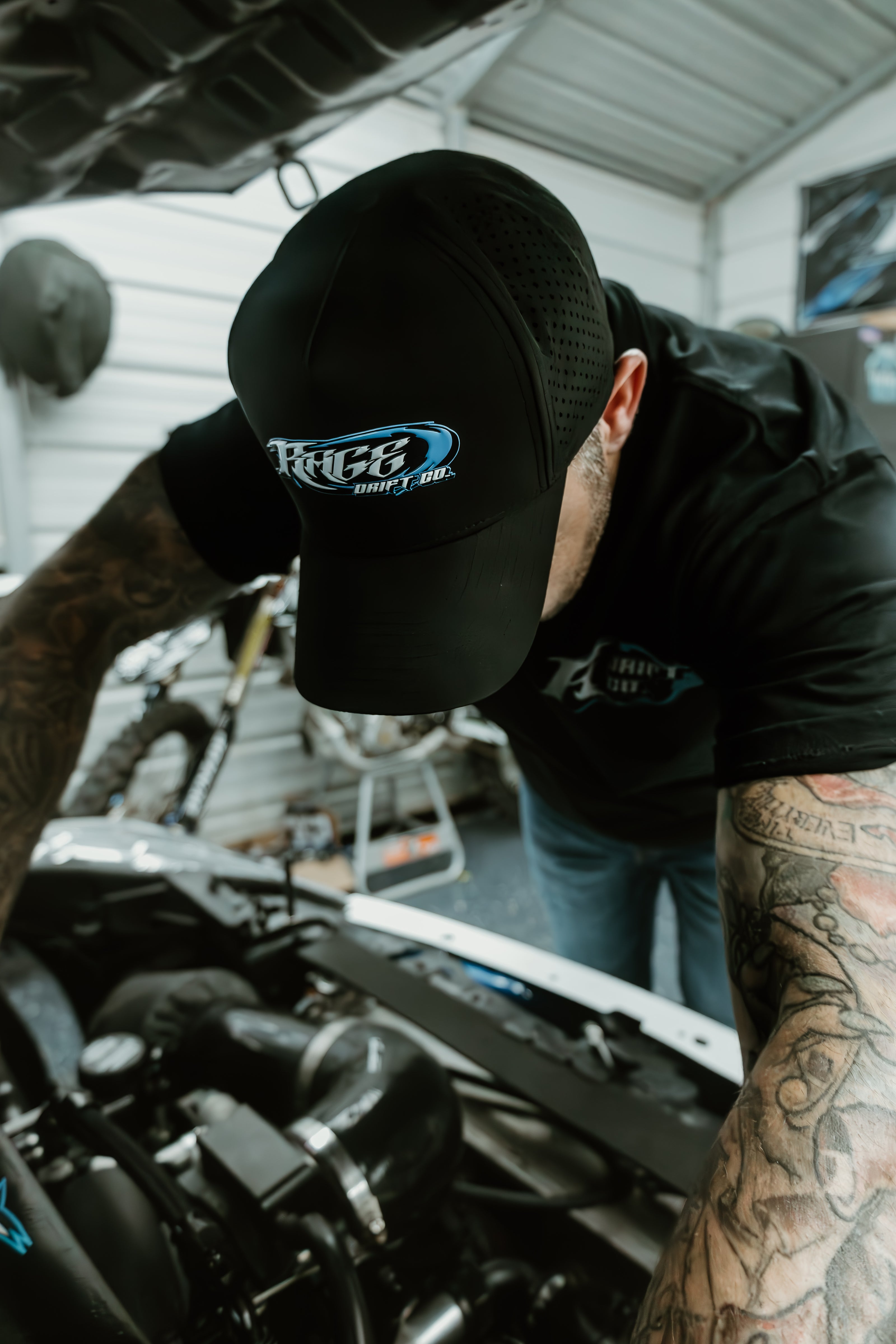 A tattooed man in a black RACE3 Racing Team cap and black shirt leans over an engine in a garage setting.