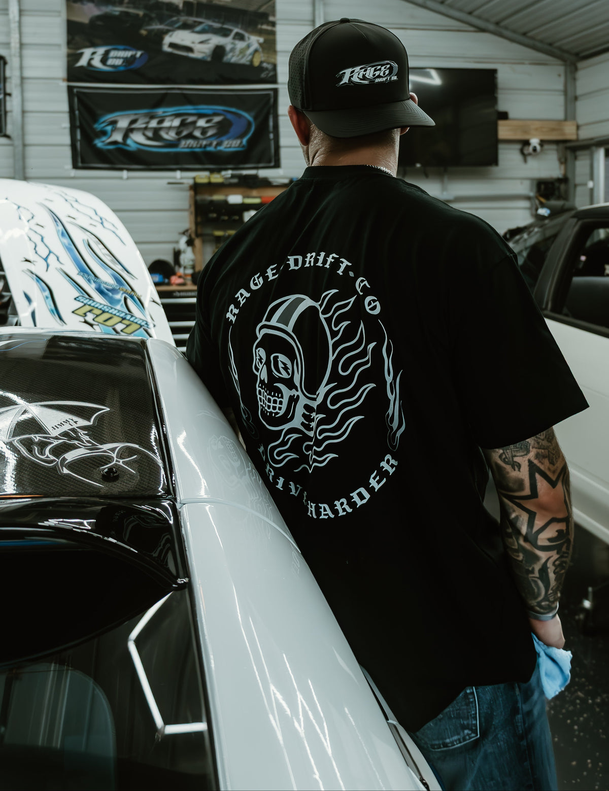 A man standing in a garage wearing a black t-shirt with a large white graphic on the back that reads 'HAGE DRIFT CO' and 'RUNNER'.