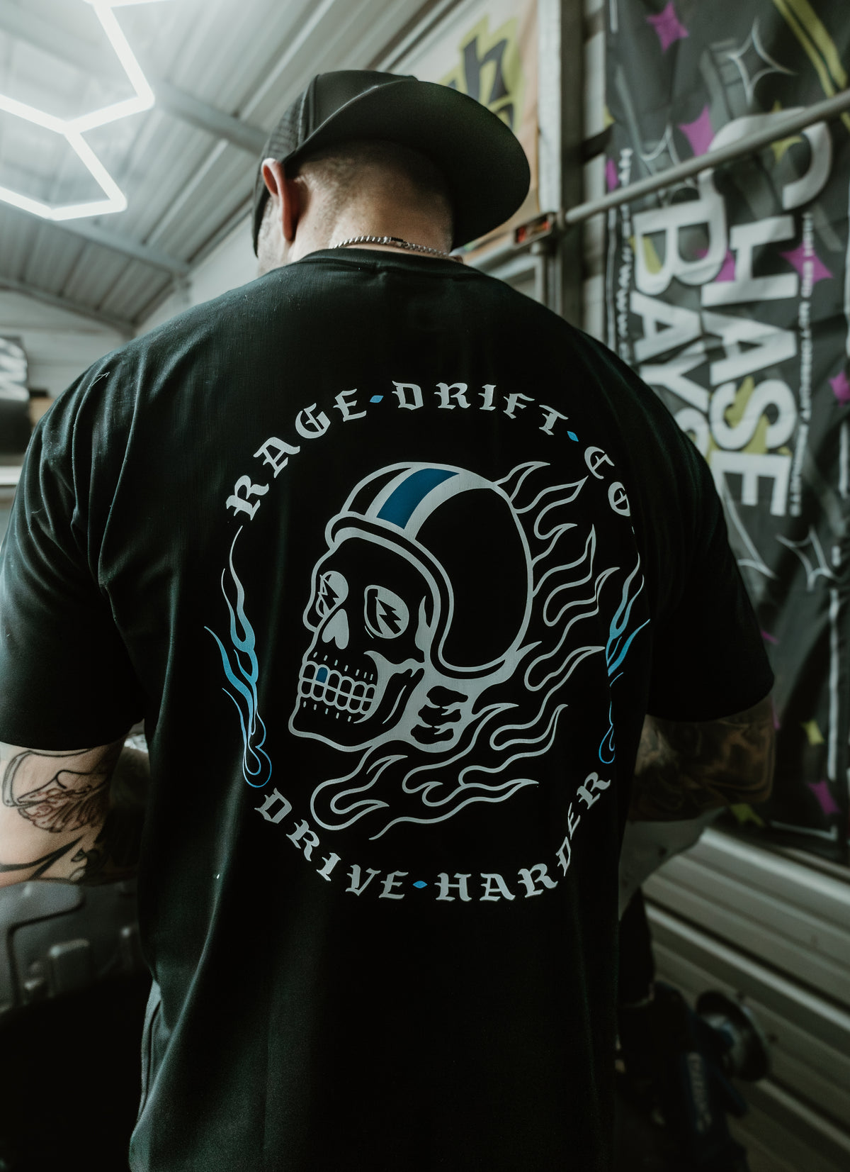 A man viewed from behind wearing a black t-shirt with a large graphic on the back. The graphic features a white and blue skull wearing a racing helmet, surrounded by the text 'RAGE DRIFT CO' and 'DRIVE HARSH'.