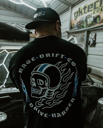 A man viewed from behind wearing a black t-shirt with a large graphic on the back, featuring a skull in a racing helmet with flames and the text 'RAGE • DRIFT • CO' and 'DRIVE • HARDER'. He is standing in a garage or workshop environment with a 'gktech.