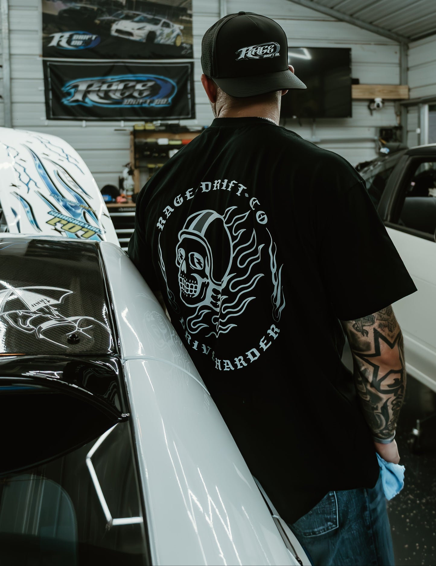 A man standing in a garage wearing a black t-shirt with a large white graphic on the back that reads 'HAGE DRIFT CO' and 'RUNNER'.