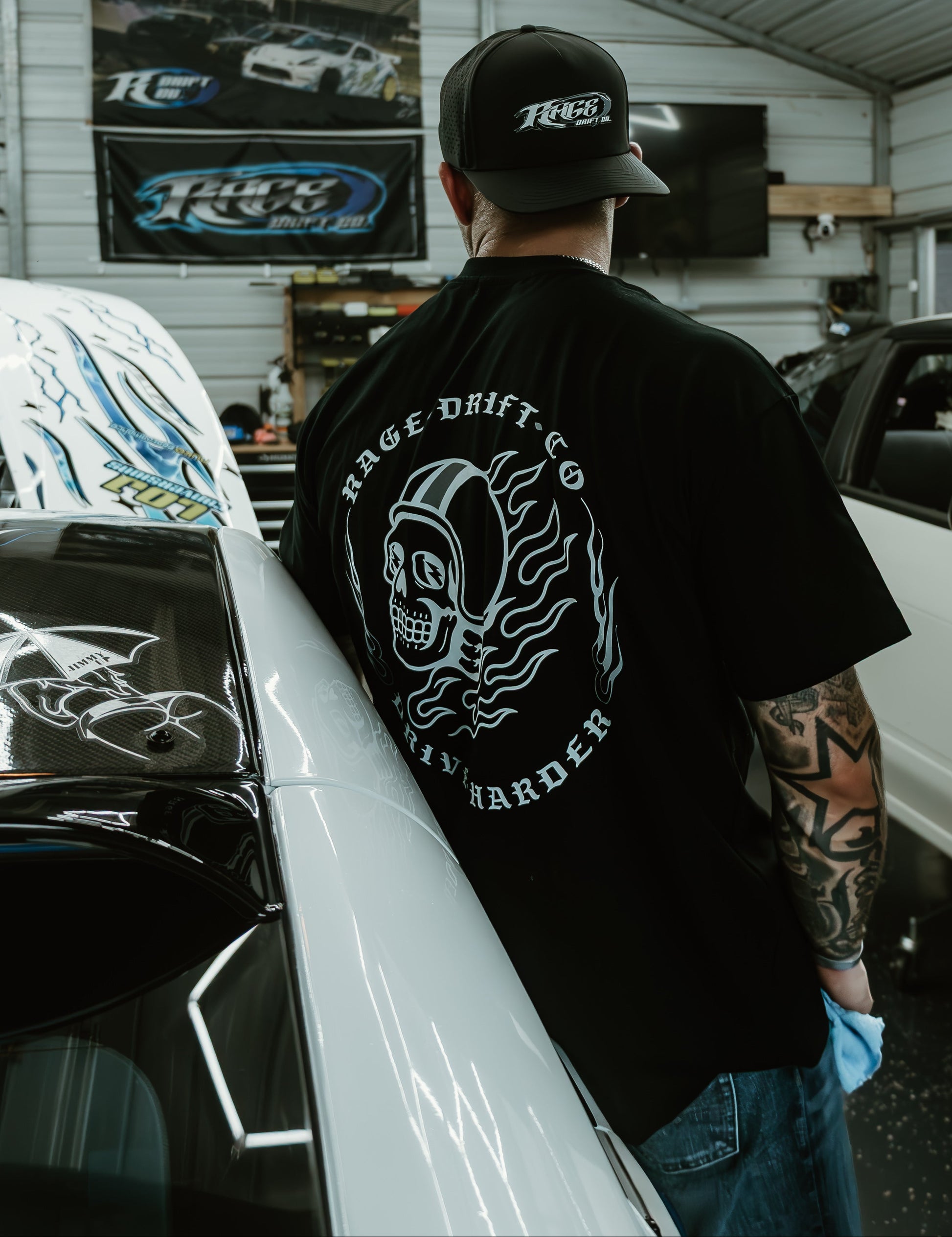A man standing in a garage wearing a black t-shirt with a large white graphic on the back that reads 'HAGE DRIFT CO' and 'RUNNER'.