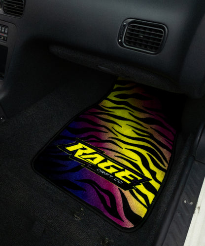 A custom car floor mat with a vibrant zebra stripe pattern in a rainbow gradient of yellow, green, blue, purple, and pink, featuring a bold black and yellow 'KZRGE' logo. The mat is shown installed inside a black car footwell.