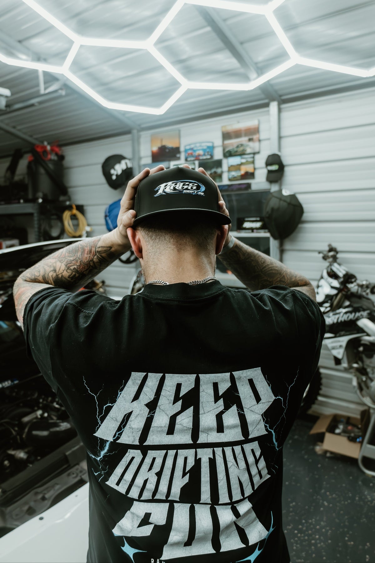 A man seen from behind wearing a black t-shirt with a large white graphic reading 'KEEP DRIFTING COOL' and a black cap, standing in a garage with motorcycle parts and hexagonal ceiling lights.