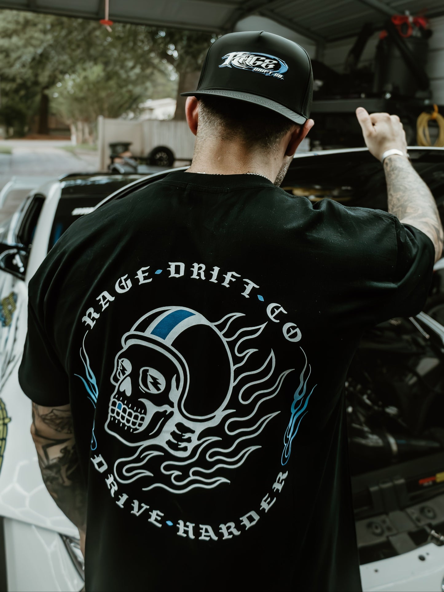 A man viewed from behind wearing a black t-shirt with a large white and blue skull graphic and the text 'RAGE DRIFT CO' and 'DRIVE HARDER', along with a black cap. He is standing in a garage next to an open car hood.